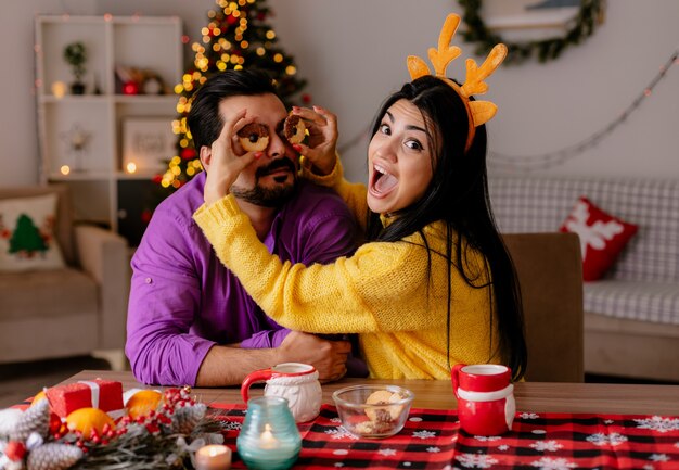 Jovem e linda casal homem e mulher sentados à mesa com biscoitos se divertindo juntos, felizes e apaixonados em um quarto decorado com árvore de natal ao fundo