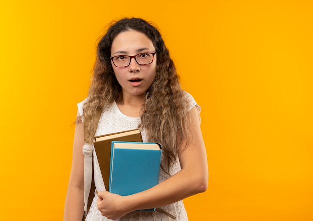 Jovem e linda colegial impressionada usando óculos e bolsa traseira segurando livros isolados em amarelo