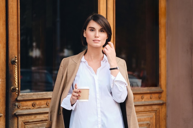 Jovem e linda empresária elegante com café para ir sonhadoramente olhando para a câmera ao ar livre