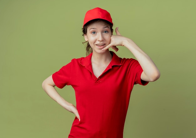Jovem e linda garota de entrega impressionada de uniforme vermelho e boné, colocando a mão na cintura e fazendo um gesto de chamada, isolado em um fundo verde oliva com espaço de cópia
