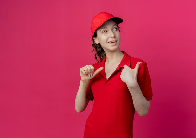 Jovem e linda garota de entrega impressionada usando uniforme vermelho e boné, olhando e apontando para o lado isolado em um fundo carmesim com espaço de cópia