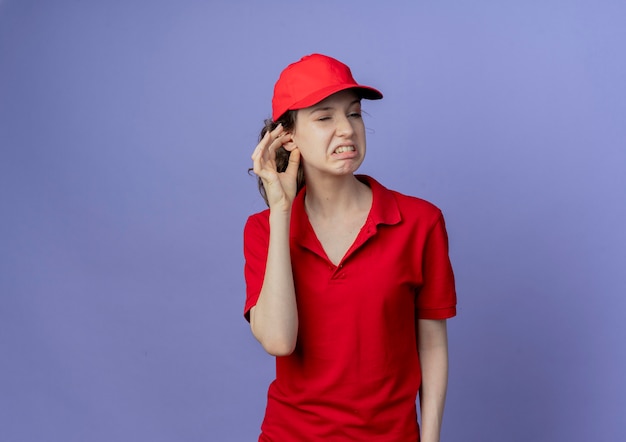 Jovem e linda garota de entrega irritada vestindo uniforme vermelho e boné olhando para o lado, colocando a mão na orelha isolada no fundo roxo com espaço de cópia