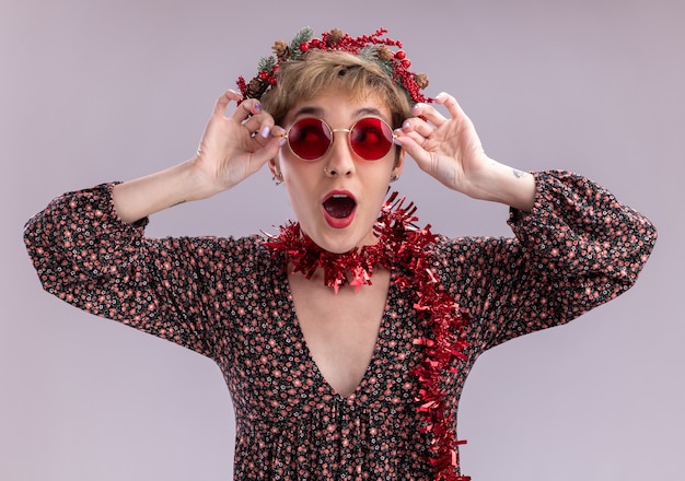 Jovem e linda garota impressionada usando coroa de flores de natal e guirlanda de ouropel em volta do pescoço com óculos olhando para o lado agarrando os óculos isolados no fundo branco