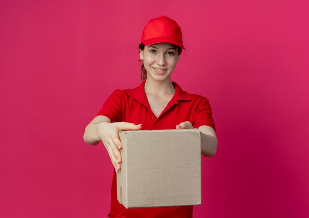 Jovem e linda garota sorridente de uniforme vermelho e boné esticando a caixa de papelão para a câmera, isolada em um fundo carmesim com espaço de cópia