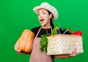 Foto grátis jovem e linda jardineira de avental e chapéu segurando uma caixa cheia de vegetais e abóbora feliz e animada