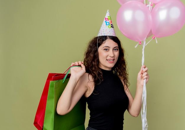 Jovem e linda mulher com cabelo encaracolado em um boné de férias segurando balões de ar e presentes de aniversário felizes e positivos sorrindo alegremente em pé sobre a parede de luz