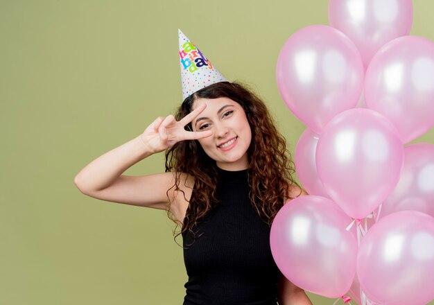Jovem e linda mulher com cabelo encaracolado em um boné de férias segurando balões de ar, feliz e positiva, mostrando o sinal V comemorando a festa de aniversário em pé sobre a parede de luz