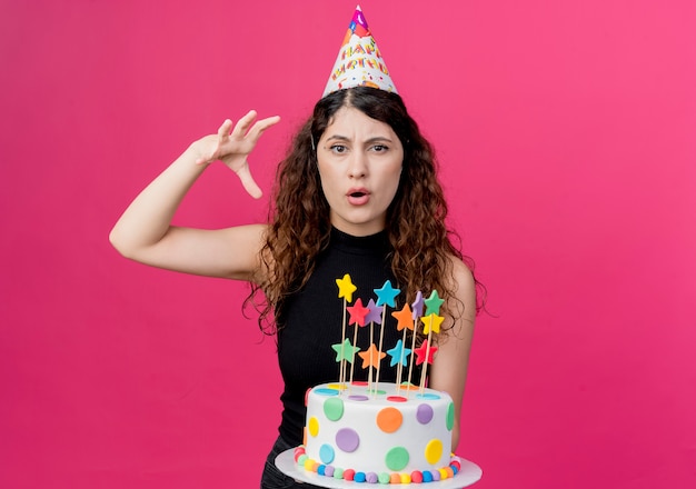Jovem e linda mulher com cabelo encaracolado em um boné de férias segurando um bolo de aniversário descontente com o conceito de festa de aniversário em pé sobre a parede rosa