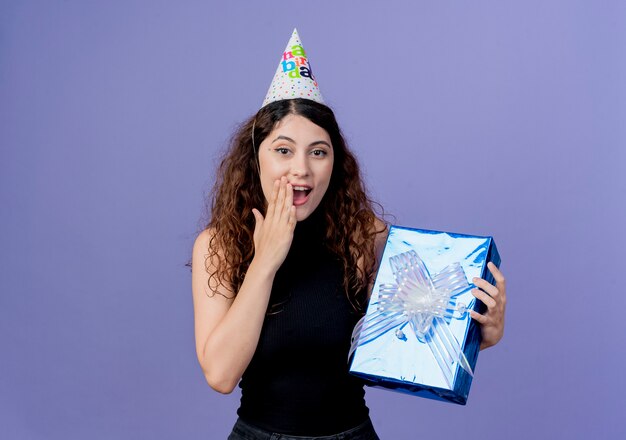 Jovem e linda mulher com cabelo encaracolado em um boné de férias segurando uma caixa de presente de aniversário e parecendo espantada com o conceito de festa de aniversário em pé sobre a parede azul