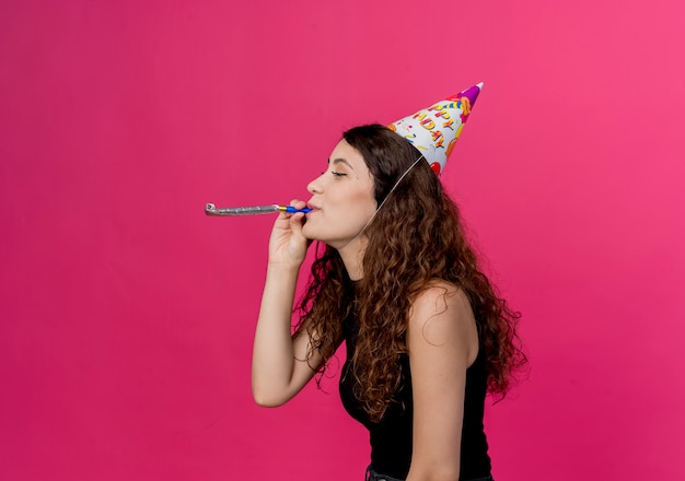 Foto grátis jovem e linda mulher com cabelo encaracolado em um boné de férias soprando apito conceito de festa de aniversário em pé sobre a parede rosa