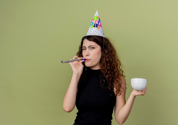 Jovem e linda mulher com cabelo encaracolado em um boné de férias, soprando apito segurando a xícara de café, parecendo displicente com o conceito de festa de aniversário sobre a luz