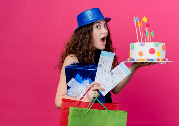 Jovem e linda mulher com cabelo encaracolado em um chapéu de férias segurando uma caixa de presente de bolo de aniversário e passagens aéreas conceito de festa de aniversário feliz e animado em pé sobre a parede rosa