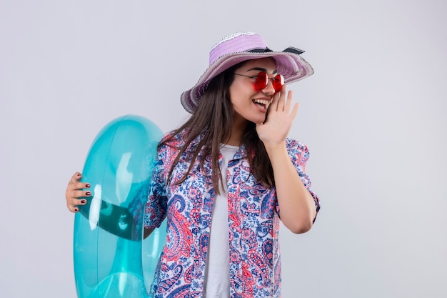 Jovem e linda mulher com chapéu de verão e óculos de sol vermelhos segurando um anel inflável, gritando ou chamando alguém com a mão perto da boca positiva e feliz, olhando de lado pronto para o conceito de férias