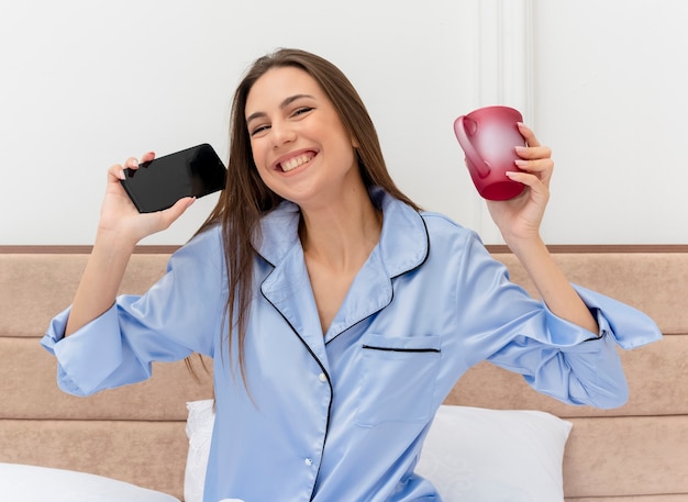 Jovem e linda mulher de pijama azul sentada na cama com uma xícara de café segurando o smartphone olhando para a câmera feliz e animada sorrindo alegremente no interior do quarto sobre fundo claro