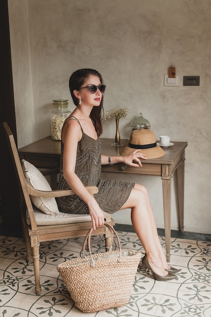 Jovem e linda mulher elegante em um quarto de hotel de resort, sentada à mesa, usando um vestido da moda, estilo safári, chapéu de palha, sorrindo, feliz, férias de verão, roupa boêmia, bolsa de praia, óculos de sol, pernas