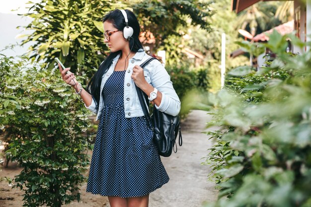 Jovem e linda mulher elegante usando smartphone, fones de ouvido, óculos, verão, roupa jeans vintage, sorrindo, feliz, positiva