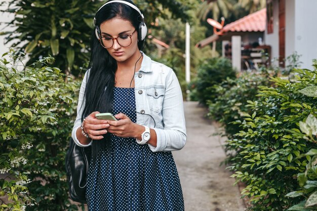 Jovem e linda mulher elegante usando smartphone, fones de ouvido, óculos, verão, roupa jeans vintage, sorrindo, feliz, positiva