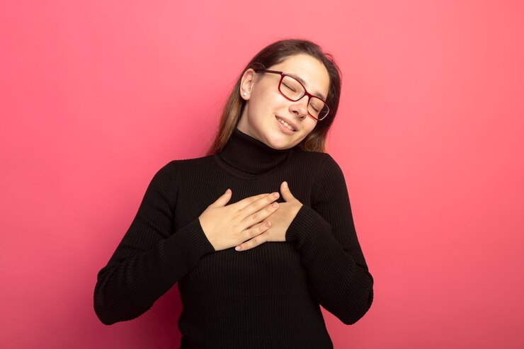 Foto grátis jovem e linda mulher em uma blusa de gola alta preta e óculos olhando para a frente de mãos dadas no peito, sentindo emoções positivas com os olhos fechados, em pé sobre a parede rosa
