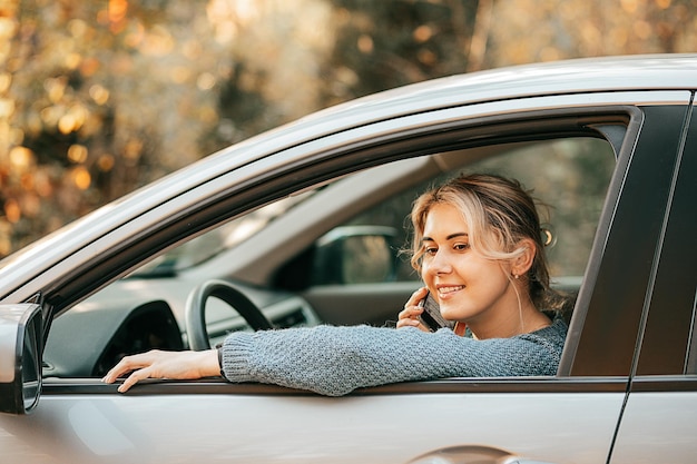 Jovem e linda mulher loira falando na janela do carro do smartphone aberta enquanto para o veículo sorrindo