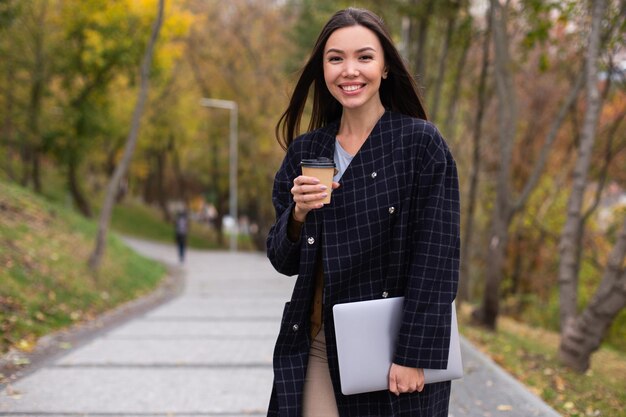 Jovem e linda mulher sorridente de casaco com café para ir e laptop olhando alegremente para a câmera no parque outono