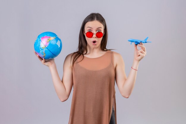 Jovem e linda mulher usando óculos escuros vermelhos segurando um globo e um avião de brinquedo espantada e surpresa em pé sobre um fundo branco