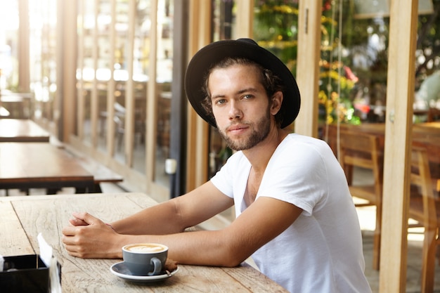 Jovem elegante com a barba por fazer e um olhar alegre, sentado à mesa de madeira em um café ao ar livre com uma xícara de cappuccino na frente dele