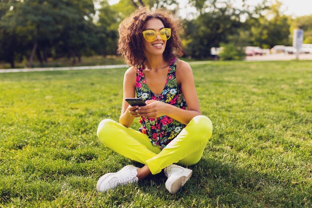 Jovem elegante e sorridente mulher negra usando smartphone, ouvindo música em fones de ouvido sem fio, se divertindo no parque, estilo colorido da moda do verão, sentada na grama, óculos de sol amarelos