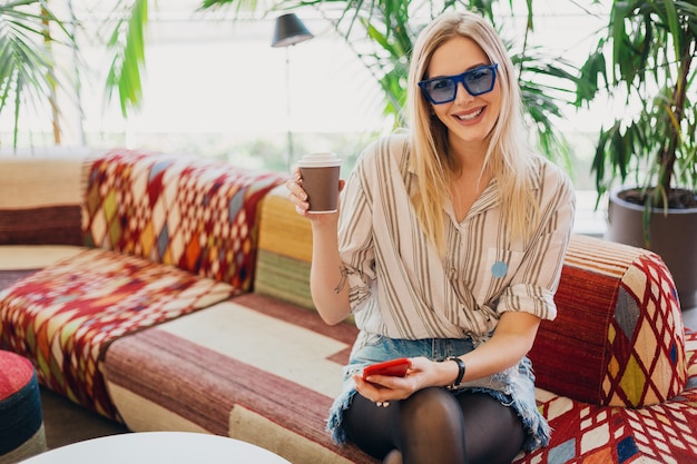 Jovem elegante e sorridente, sentada em um café moderno no sofá, vestindo camiseta e óculos de sol azuis, bebendo café