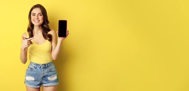 Foto grátis jovem elegante mostrando a tela do celular apontando para a interface do aplicativo de exibição do smartphone ou loja online sorrindo satisfeito em pé sobre fundo amarelo