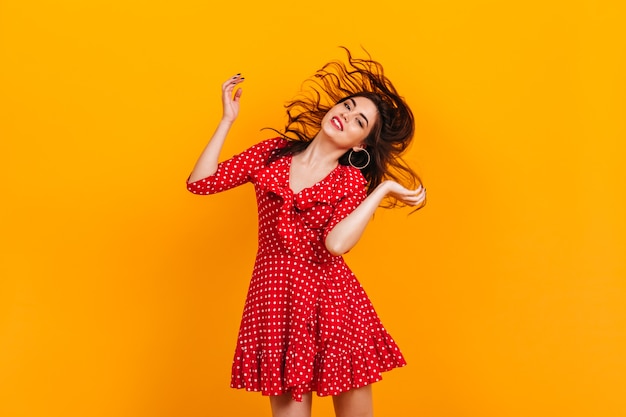 Jovem elegante num vestido curto vermelho joga o cabelo. retrato de morena em brincos de argola na parede amarela.