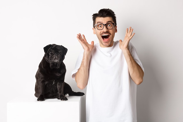 Jovem em êxtase olhando com entusiasmo e alegria, parado perto de um lindo pug preto, olhando feliz para a câmera, em pé sobre um fundo branco