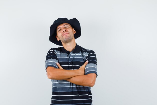 Jovem em pé com os braços cruzados em t-shirt, chapéu e olhando confiante, vista frontal.