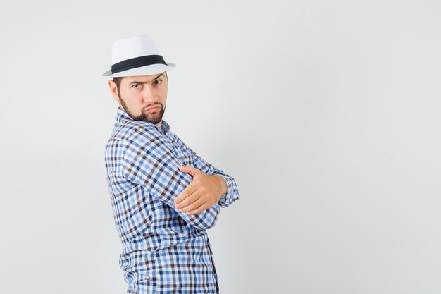 Jovem em pé com os braços cruzados na camisa, chapéu e olhando sério.