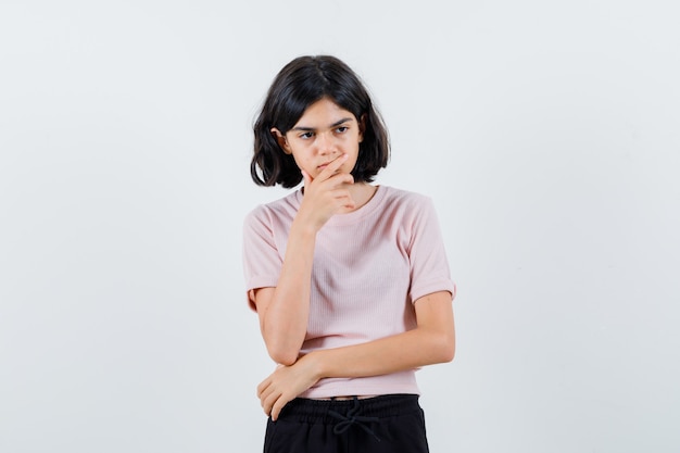 Jovem em pose pensativa, colocando a mão no queixo em uma camiseta rosa e calça preta e parecendo pensativa