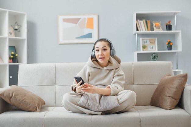 Jovem em roupas casuais com fones de ouvido sentado em um sofá no interior de casa usando smartphone olhando para a câmera feliz e alegre sorrindo alegremente aproveitando seu tempo