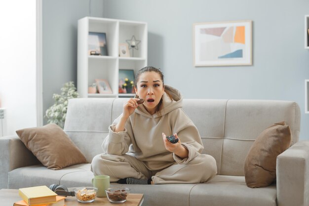 Jovem em roupas para casa, sentado em um sofá em casa comendo biscoito segurando remoto assistindo televisão sendo espantado e surpreso