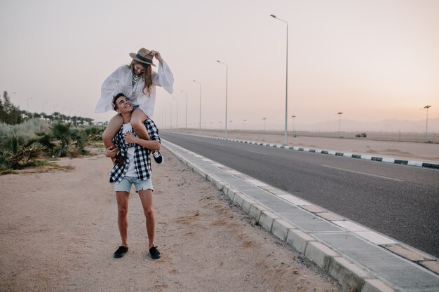 Jovem em shorts jeans, segurando sua graciosa namorada nos ombros, em pé perto da rodovia. Adorável mulher com blusa branca vintage passando um tempo com o namorado e se divertindo em um encontro ao ar livre