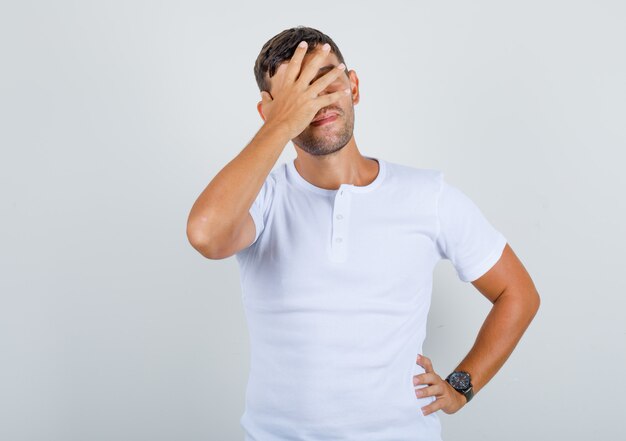 Jovem em t-shirt branca cobrindo os olhos com a mão na cintura e olhando positiva, vista frontal.