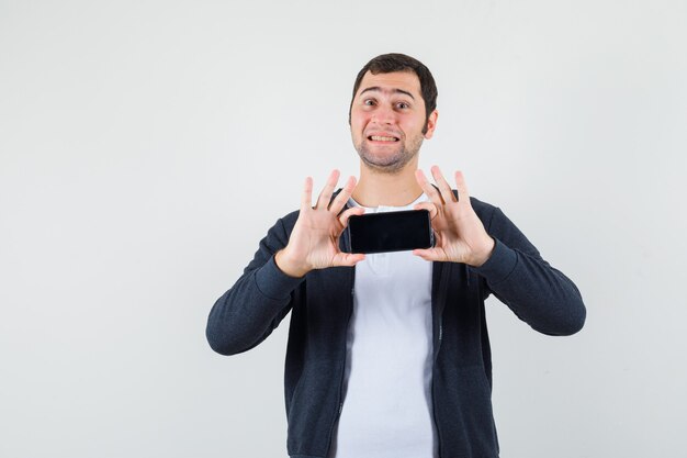 Jovem em t-shirt branca e capuz preto com zíper na frente segurando o smartphone com as duas mãos e olhando feliz, vista frontal.