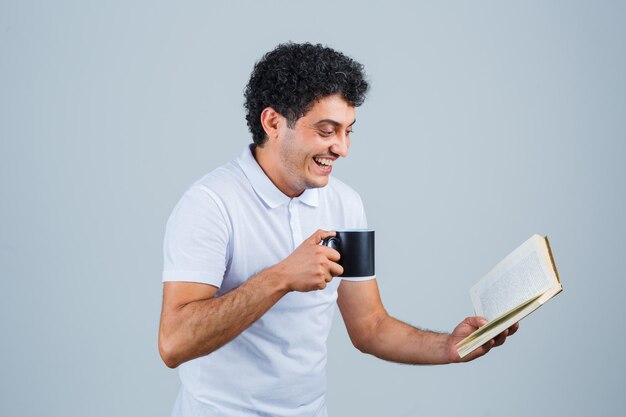 Jovem em t-shirt branca e jeans, bebendo chá enquanto lê um livro e parece feliz, vista frontal.