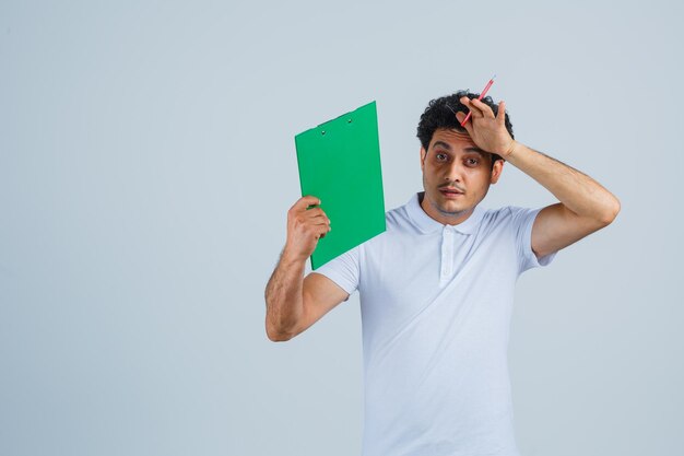 Jovem em t-shirt branca e jeans, segurando o caderno e uma caneta, segurando a mão na testa e parecendo estressado, vista frontal.