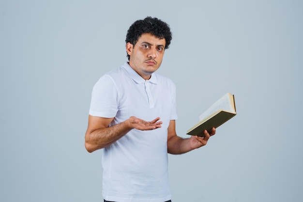 Jovem em t-shirt branca e jeans segurando o livro e esticando a mão de forma questionadora e parecendo perplexo, vista frontal.