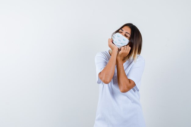 Jovem em t-shirt branca e máscara repousando o rosto nas mãos e olhando com sono, vista frontal.