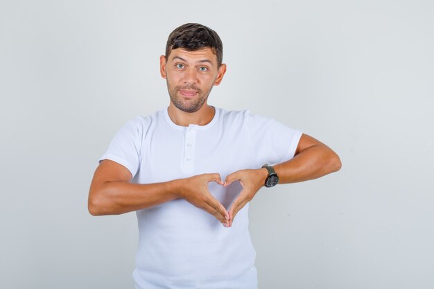 Jovem em t-shirt branca fazendo um gesto de coração com os dedos e olhando positiva, vista frontal.