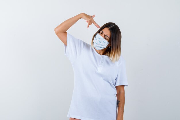 Jovem em t-shirt branca, máscara mostrando gesto de arma perto da cabeça e olhando confiante, vista frontal.