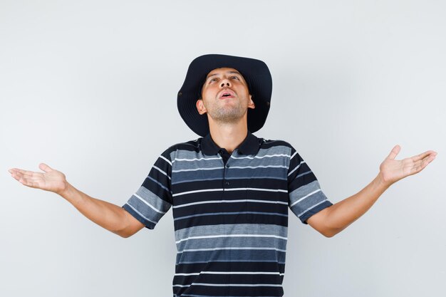 Jovem em t-shirt, chapéu abrindo os braços enquanto olha para cima e parece agradecido, vista frontal.