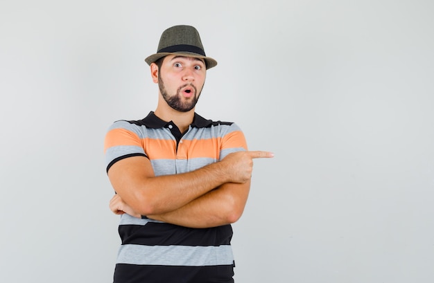 Foto grátis jovem em t-shirt, chapéu apontando para o lado e olhando curioso, vista frontal.