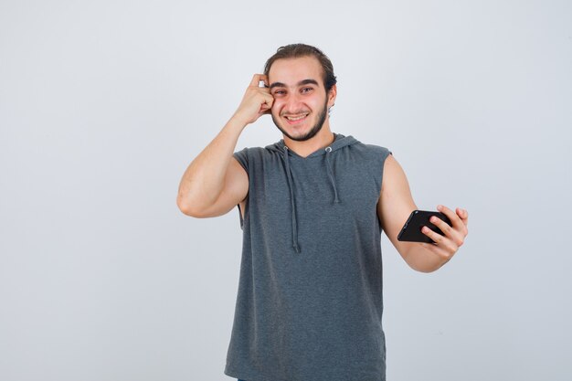 Jovem em t-shirt com capuz, segurando o telefone na mão, segurando a mão na cabeça e olhando alegre, vista frontal.