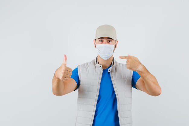 Jovem em t-shirt, jaqueta, boné, apontando para a máscara, aparecendo o polegar para cima, vista frontal.