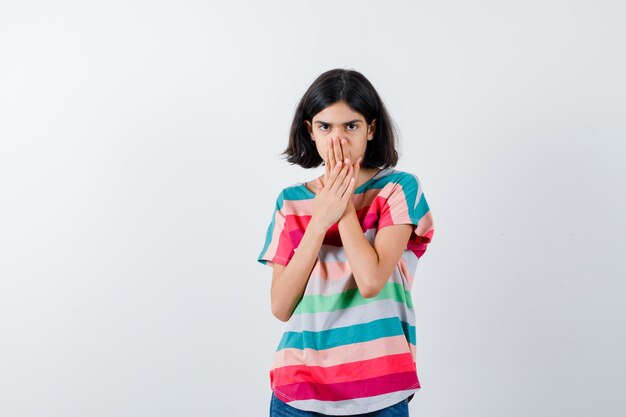 Jovem em t-shirt listrada colorida, cobrindo a boca com as mãos e parecendo tímida, vista frontal.